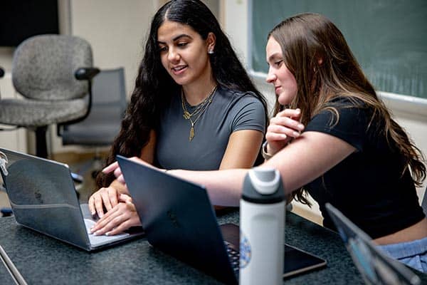 Two students work together on their laptops while sitting in a campus classroom