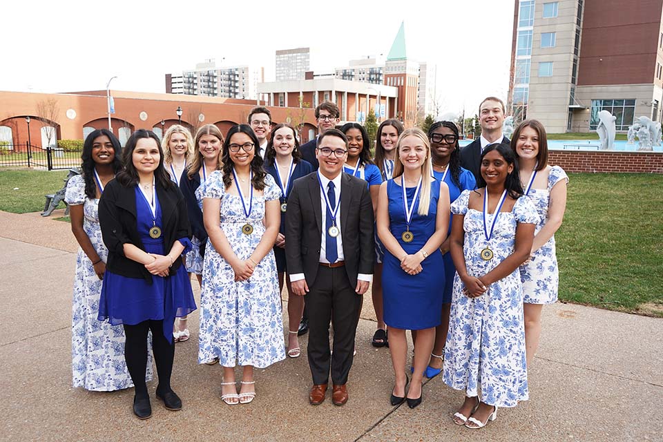 A large group of students dressed nicely and with medals around their neck pose for a photo outside on SLU's campus.