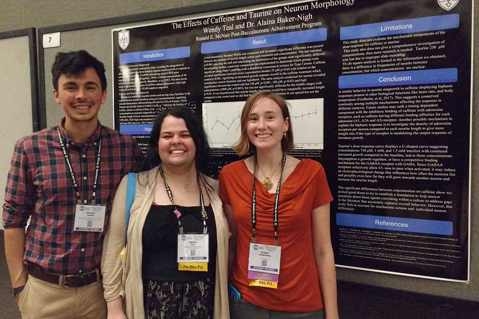 Neuroscience Three students wearing conference lanyards stand in front of a conference poster that reads "The Effects of Caffeine and Taurine on Neuron Morphology"