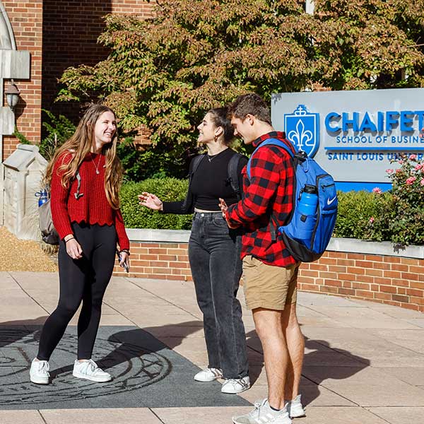 A group of three students stand and chat in front of the Chaifetz School of Business sign in front of Cook Hall.