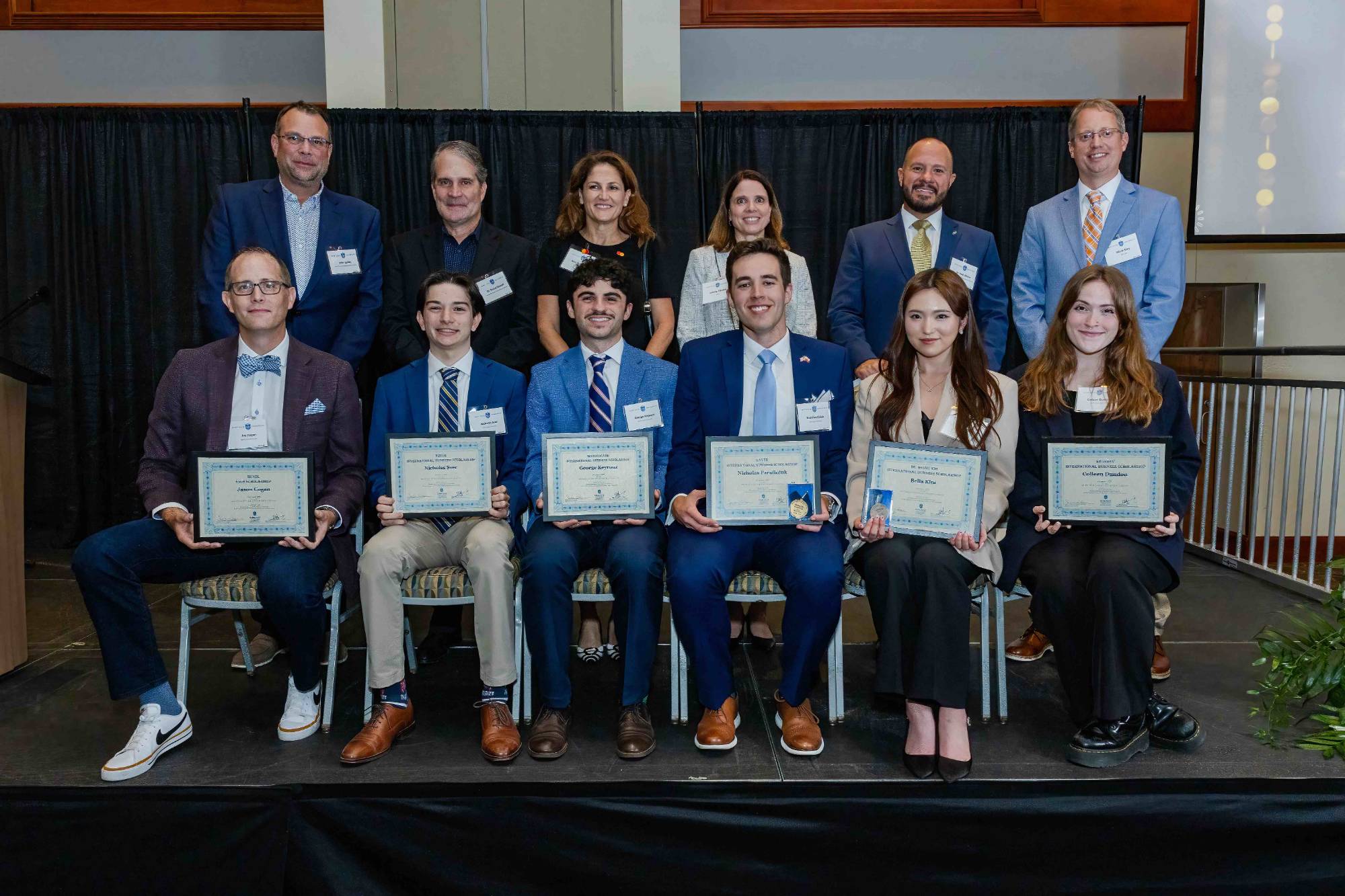 Award winners sit in a row holding up awards plaques while faculty stand behind them.
