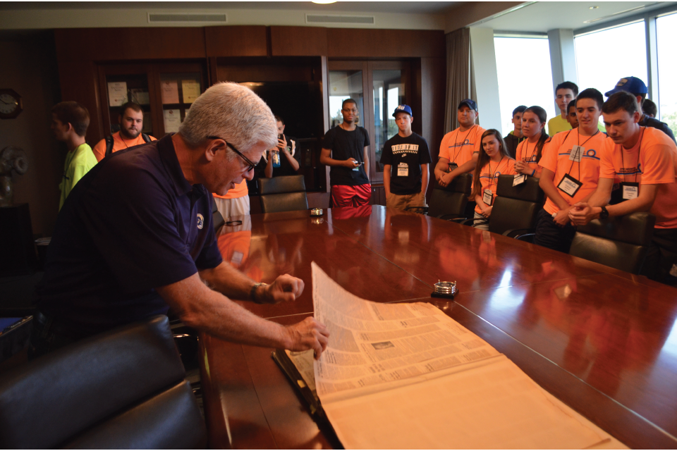 Allsup Entrepeneurship Academy Students in orange standing around a table in a conference room as part of Allsup Entrepeneurship Academy
