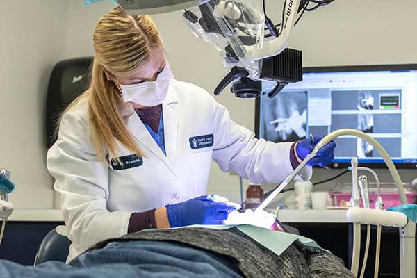 A female CADE resident, wearing a white lab coat and mask, uses a dental tool to work on the teeth of a reclining patient in a dental chair.
