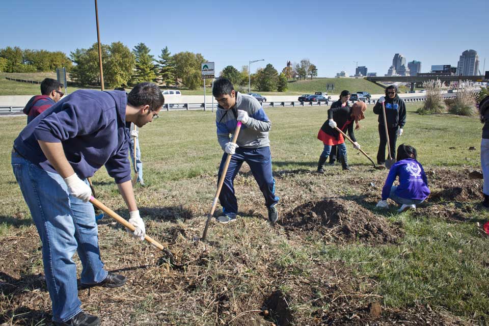 Students garden as part of a service project Students use shovels and other equipment in a grassy area next to a highway.