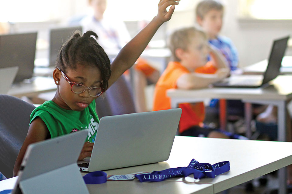 Prep Workshops A child looks at a laptop screen and raises her hand while sitting in a classroom with other students her age.