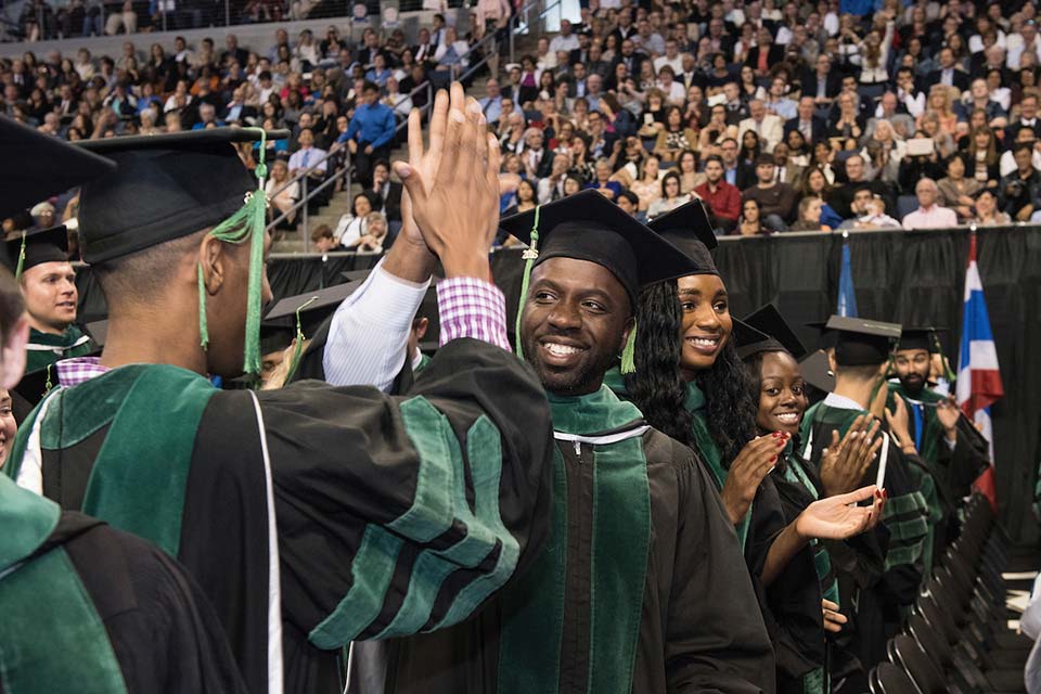 Retention Two students wearing caps and gowns high five each other in a crowded arena.