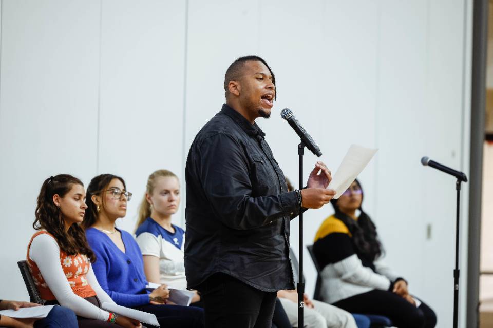 Aric Hamilton M L K speech A student stands and speaks into a microphone holding a piece of paper while other students, seated, look on