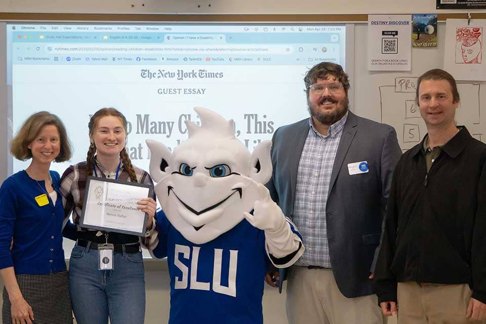 Aurora Hofius, a young female student, stands surrounded by educators and the Billiken in front of a white board.