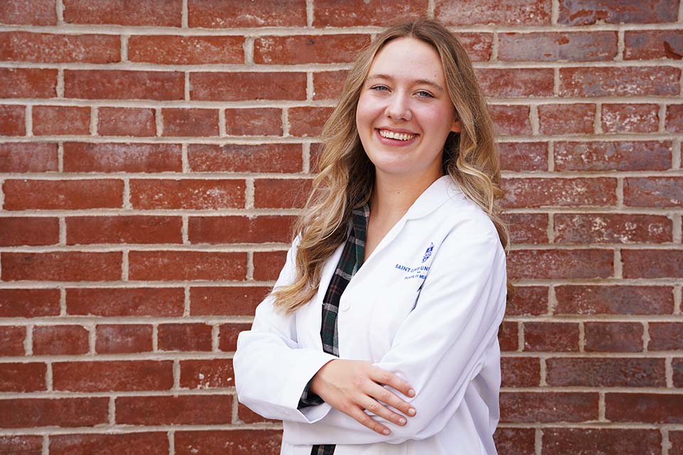 Caroline Kaczmarek Caroline poses for a photo wearing her white lab coat against a brick wall.