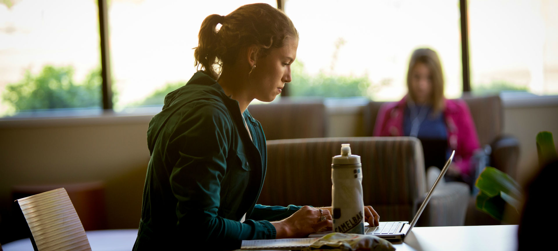 Studying A student studying at a table with a water bottle and laptop as another student sits in the background in front of a window.