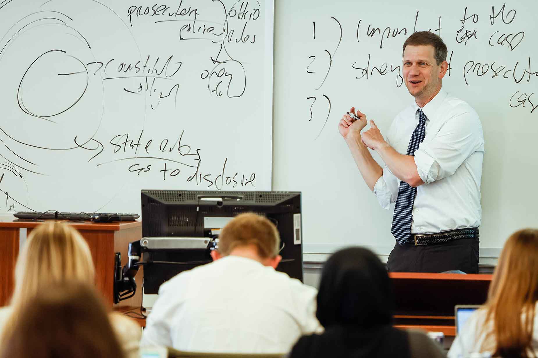 A male law professor presents a lecture to students while pointing to notes on a whiteboard during class.