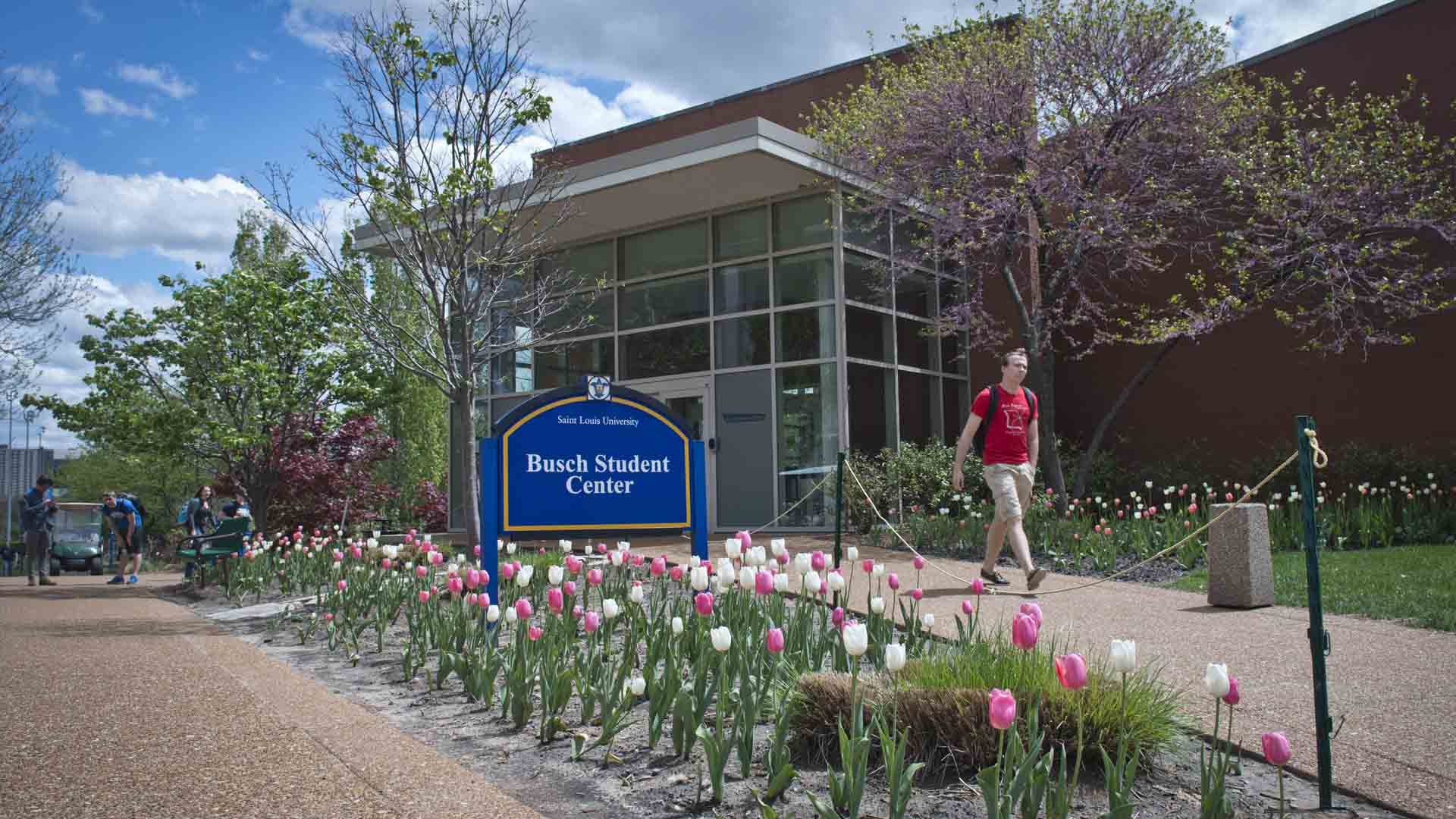 Busch Student Center Exterior of the Busch Student Center with a student walking by and flowers blooming.