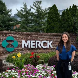 Lauren Favell standing to the right of a flower bed. Above the flower bed is a large Merck logo on a brick wall.