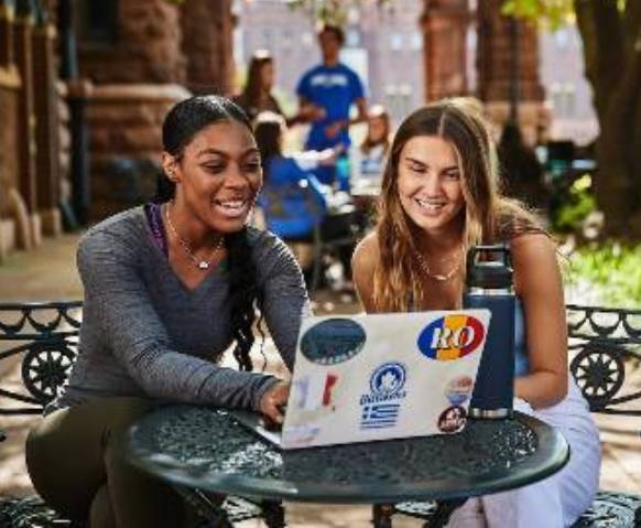 Two smiling students sitting outdoors at a small cafe table studying with laptops in front.