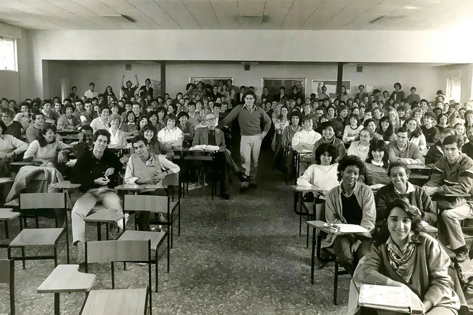 SLU-Madrid old class A black and white photo features students sitting at classroom desks while a professor stands in the center of the room.