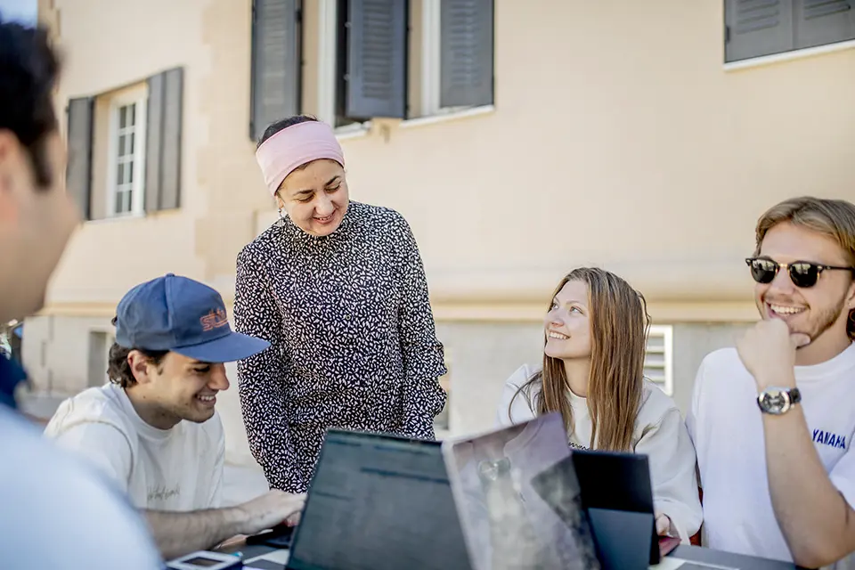 Computer Science class Students sit with laptops at outdoor tables, holding a discussion with a professor.