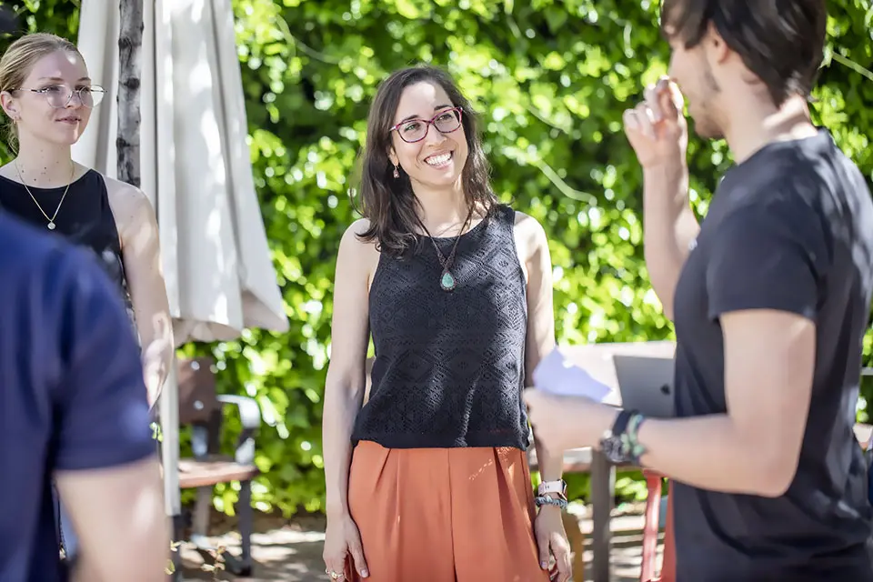 Humanities and Social Sciences Students converse while standing outside under some trees on a sunny day.
