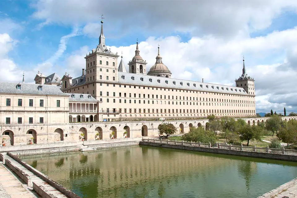 The Escorial Monastery Exterior shot of a monastery with a large pond in the patio. The building is made from light bricks and contains multiple arches on the ground floor, with several floors containing rows of windows.