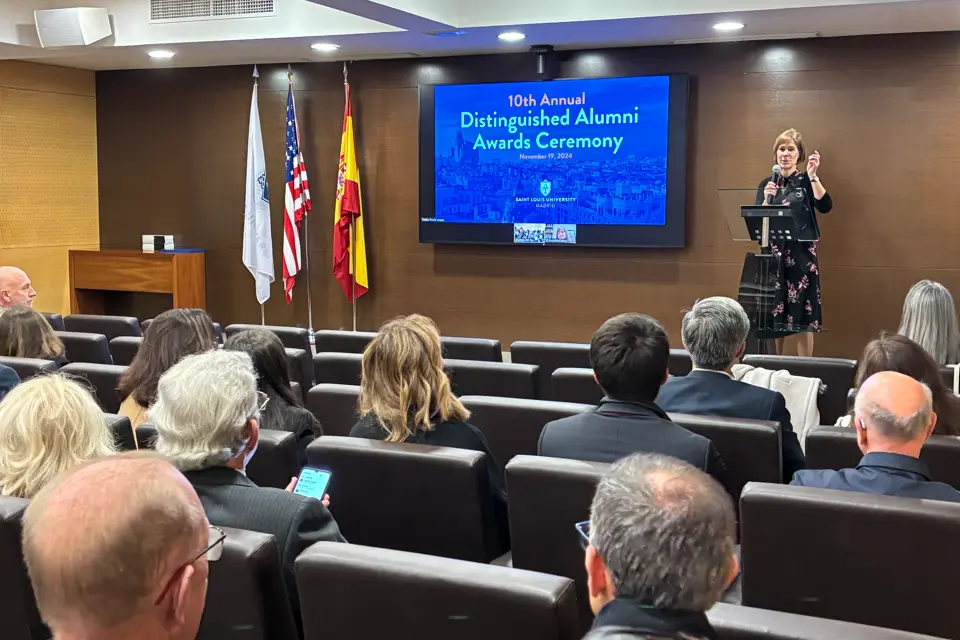 SLU-Madrid Celebrates 10th Annual Alumni Awards Woman with a microphone standing behind a lectern welcomes the audience in the auditorium with the alumni ceremony poster on screen.