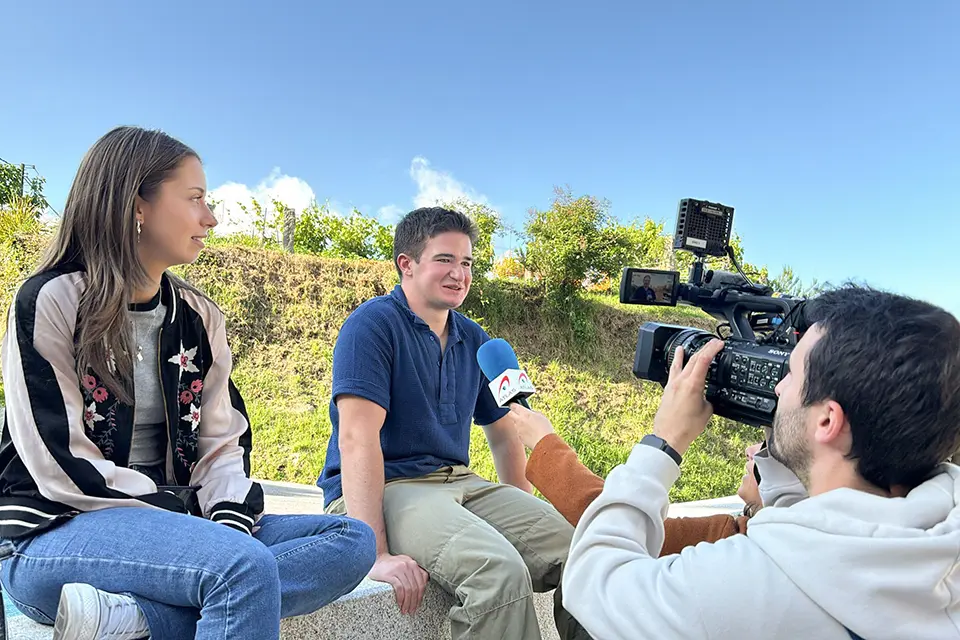 Two students sitting on a ledge while being interviewed by press that are holding a microphone and camera
