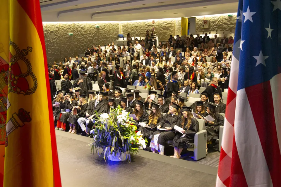 Spanish and American flags displayed at May 2023 Commencement Ceremony. Spanish and U.S. flags frame an image of graduates and sitting in an auditorium wearing caps and gowns and their families.