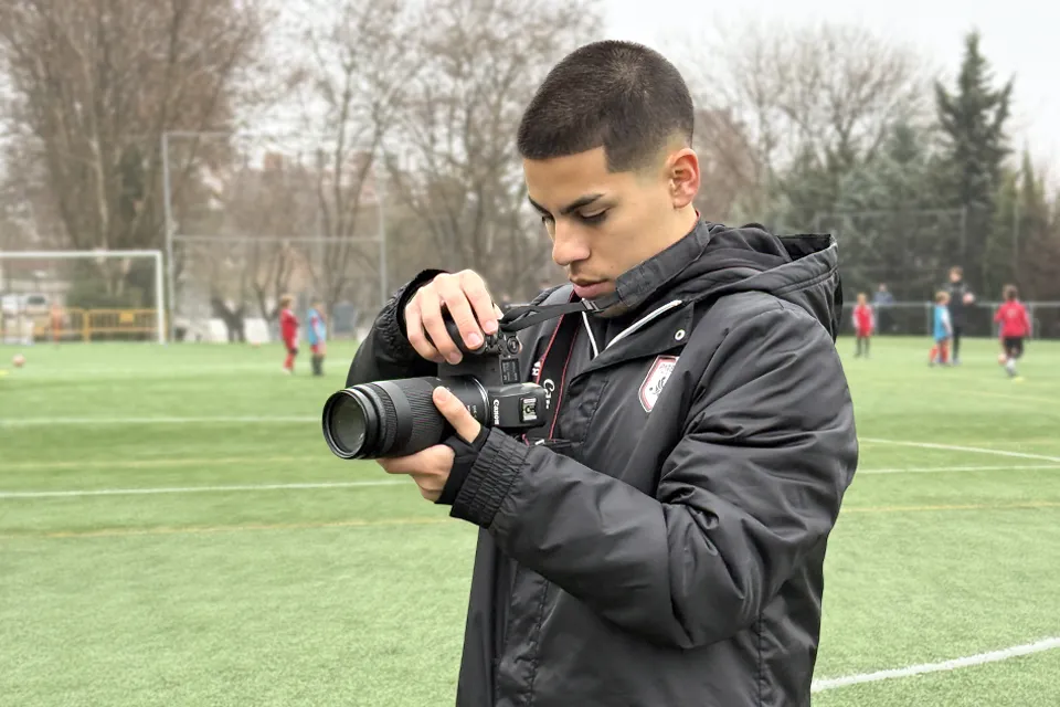 Carlos Lindo drives brand growth and visibility by developing marketing strategies. A young man holds a camera on a soccer field with children playing in the background.