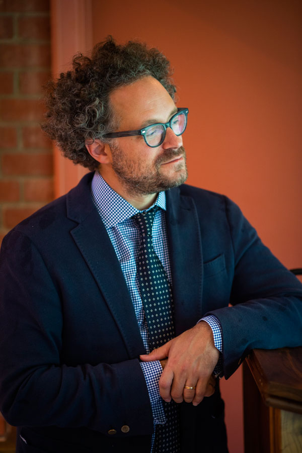 Curator Aaron Rosen, dressed in a blue suit, rests his arm on a podium and gazes to the right