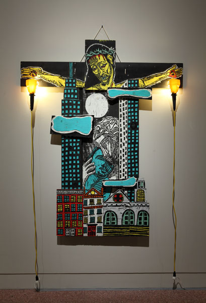 Adrian Kellard, Lovers, 1986. Latex on wood with trouble lamps. MOCRA collection. Photo by Jeffrey Vaughn. An artwork by Adrian Kellard titled Lovers. A sculptural work made up of various carved wood panels. At top, the head and arms of Jesus on the Cross. In the place of his body, a cityscape with skyscrapers and, at bottom, a street scen with apartment buildings and a church. In the middle of the work, between the skyscrapers, a man embraces another man tenderly. Yellow trouble lamps are suspended from the hands of Christ, with the power cords dangling down.