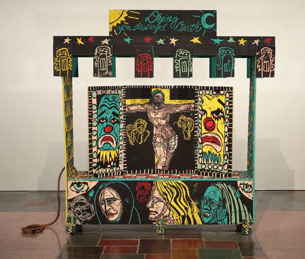 Adrian Kellard, Shrine, 1984–1985. Latex on wood with rope and hardware. MOCRA collection. Photo by Jeffrey Vaughn. An artwork by Adrian Kellard titled Shrine. A large wagon made of carved wood panels painted in yellow, teal, red, and black, features carved drawings including Jesus on the Cross, clown faces, whimsical angels, and the words, "Dying you destroyed Death."