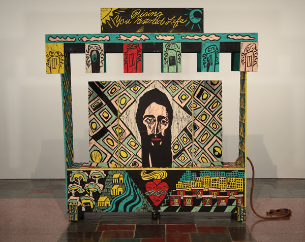 Adrian Kellard, Shrine, 1984–1985. Latex on wood with rope and hardware. MOCRA collection. Photo by Jeffrey Vaughn. An artwork by Adrian Kellard titled Shrine. A large wagon made of carved wood panels painted in yellow, teal, red, and black, features carved drawings including the face of Jesus, the Sacred Heart, buildings and a landscape, whimsical angels, and the words, "Rising You restored Life."