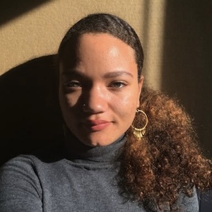 A shoulder-length photograph of Summer Sloane-Britt in a heather-gray turtleneck sweater and large gold hoop earrings. She stands against a caramel-colored background.