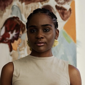 Sydney Vernon A shoulder-length photograph of a young adult woman with plaited black hair drawn into a light yellow band behind her head. She wears a sleeveless cream-colored top and stands looking slightly down at the camera with an intent gaze. In the background, out of focus, is a painting with a woman in profile who is positioned just to the right of the subject's head.