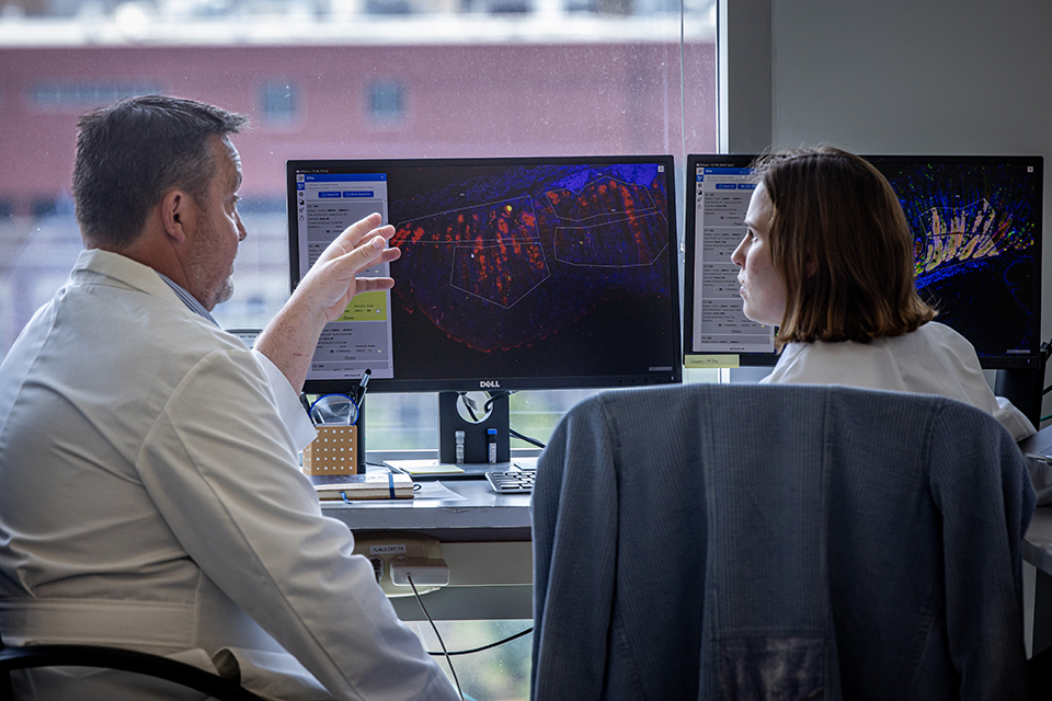 Stella Hoft, right, and Richard DiPaolo, Ph.D, discuss data on May 24, 2023. A photo of two people inside a lab wearing white coats. They sit in front of computer monitors. They discuss colorful data displayed on the screen.