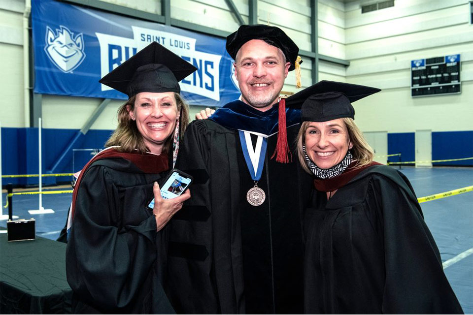 Faculty at Commencement Three professors wearing academic regalia smile while standing in a gymnasium.