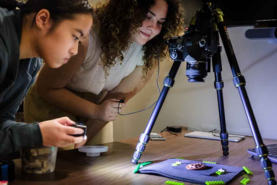 Digital Humanities Two students lean over a table to examine a round, copper-colored artifact that is placed under a camera-like device attached to a tripod.