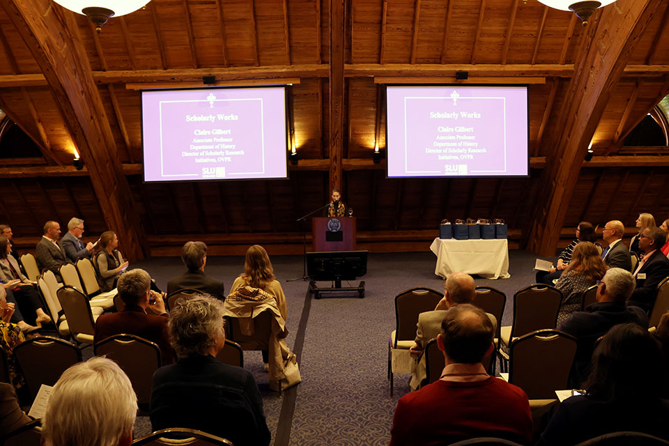 Claire Gilbert, Ph.D., speaks at the Grant Winners and Scholarly Works Reception, hosted by the Office of the Vice President for Research on April 5, 2024. A woman stands at a lectern in front of an audience, with a video screen on each side