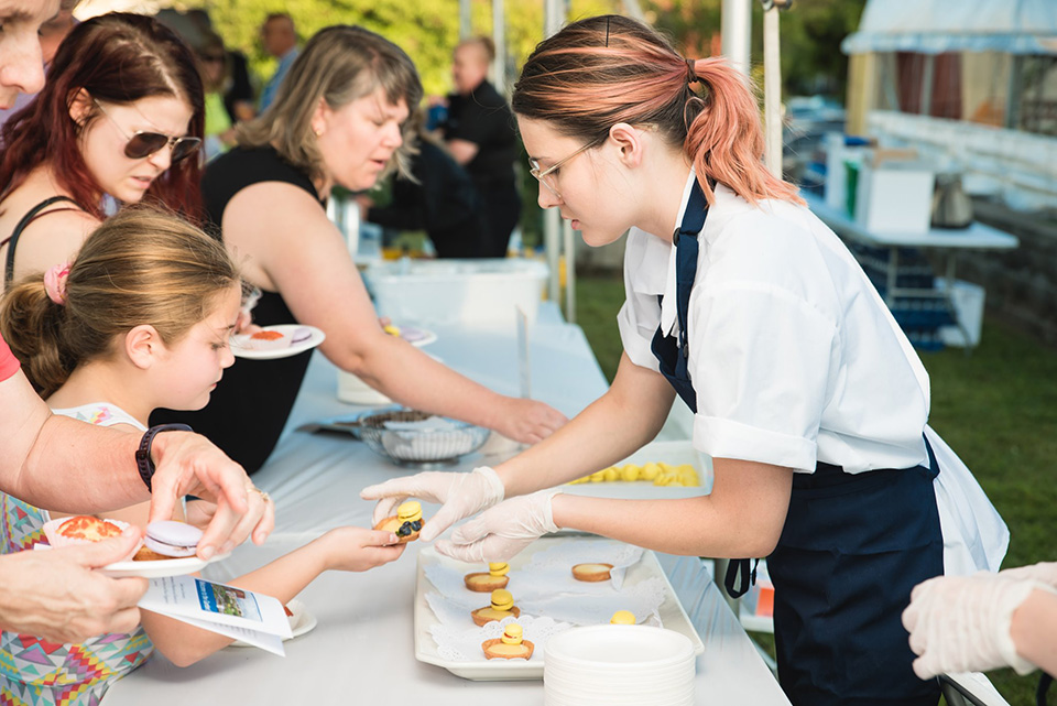 A student serving guests at the 12th Annual SLU Food Day. A student wearing an apron reaches over a table to hand a piece of food to a child.