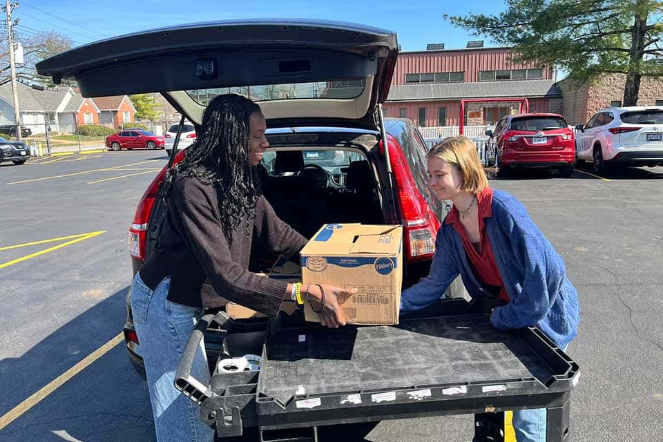 Two female volunteers load the trunk of a car with a box of food to be delivered.