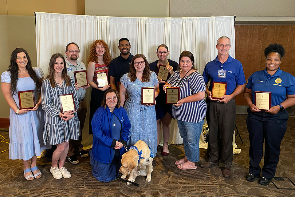 2024 Student Development Staff Excellence Award Winners A group of award winners pose for a photo, each holding a plaque. Woody, the dog, is also in the picture, but is not holding a plaque.